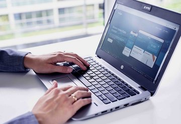 Close-up of hands typing on a laptop keyboard. The laptop screen displays the user interface of a digital application. The laptop features a VTG logo