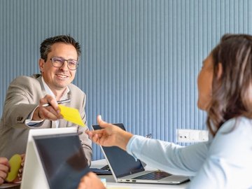 Vier Personen in einem Meeting; ein Mann reicht einer Frau einen gelben Zettel über den Tisch. Alle wirken freundlich und engagiert.